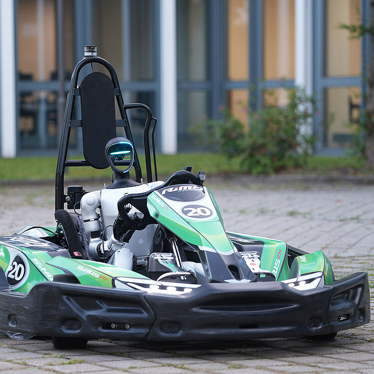Ein grünes Renn-Gokart mit der Nummer 20 steht auf einem gepflasterten Platz. Am Steuer sitzt ein humanoider Roboter mit leuchtenden blauen Augen, der das Fahrzeug autonom zu steuern scheint. 