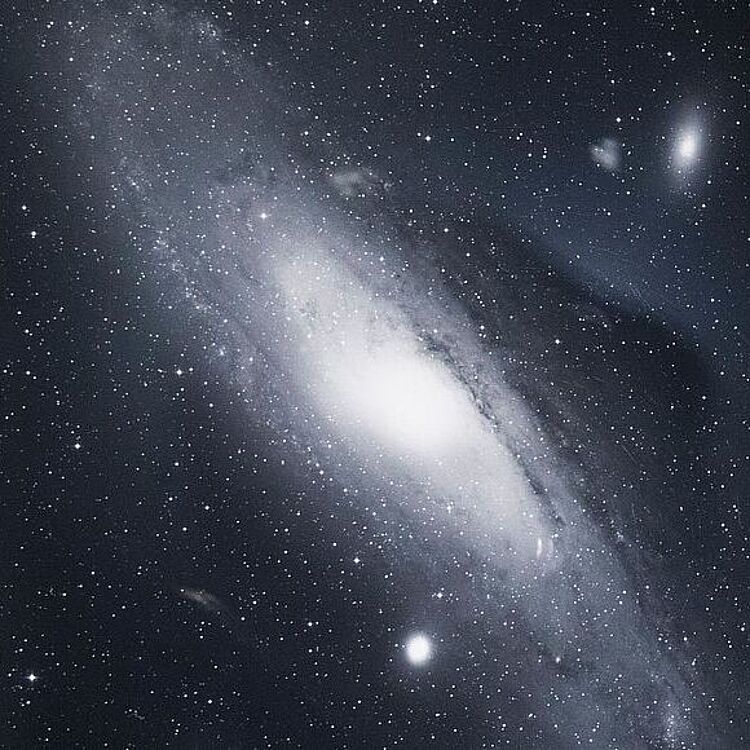 Fotografie einer Galaxie im Weltraum. Zu sehen ist eine große, spiral­förmige Ansammlung von Sternen mit hellem Zentrum und weit auslaufenden Armen, umgeben von zahlreichen kleineren Lichtpunkten, die andere Sterne und Galaxien darstellen.