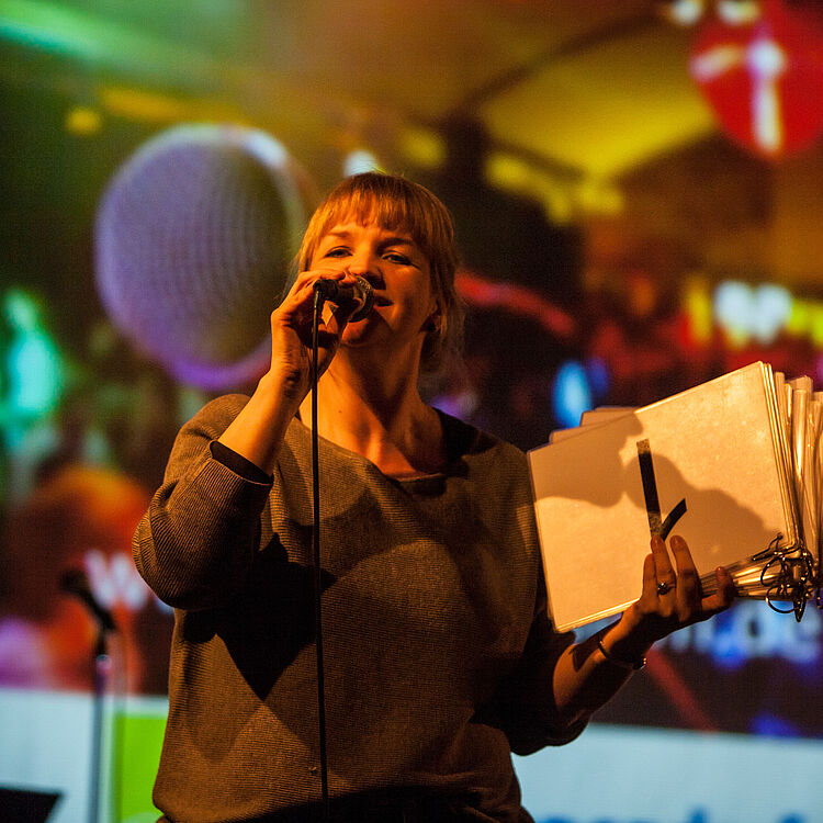 Eine auf der Bühne stehende Person, die in ein Handmikrofon spricht und mit der anderen Hand einen dicken Ringordner mit mehreren Klarsichthüllen hält; unscharfer, farbenfroher Publikum- und Lichtprojektion-Hintergrund mit warmen Gelb-, Grün- und Rottönen.