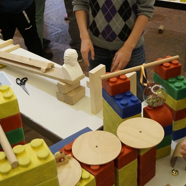 Kinder und Jugendliche bauen an einem Holzkugelbahn-Experiment: Tische mit bunten großen Steckbausteinen, Holzplatten, Rollen, Rampen und Werkzeugen in einem Innenraum; Gesichter unscharf.