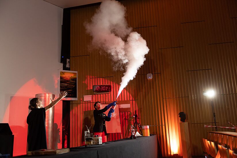Ein Student führt ein Experiment vor: Weißer Dampf schießt aus einer Flasche in die Luft. Dies freut eine Schauspielerin, die begeistert mit ihren Armen in die Luft zu dem weißen Dampf deutet.  