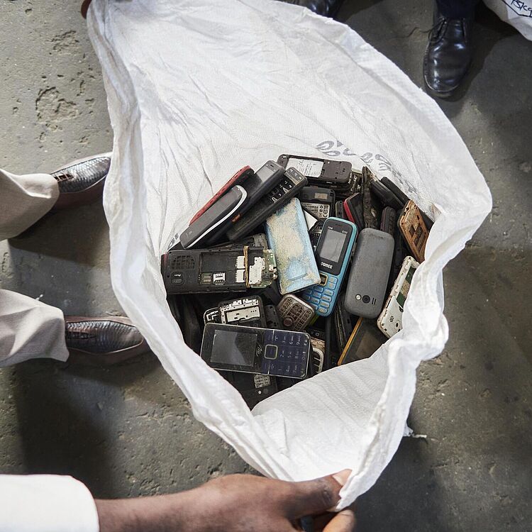 Blick von oben in einen großen weißen Sack voller ausrangierter Handys und Mobiltelefonteile, umgeben von weiteren beschrifteten Säcken auf Betonboden.