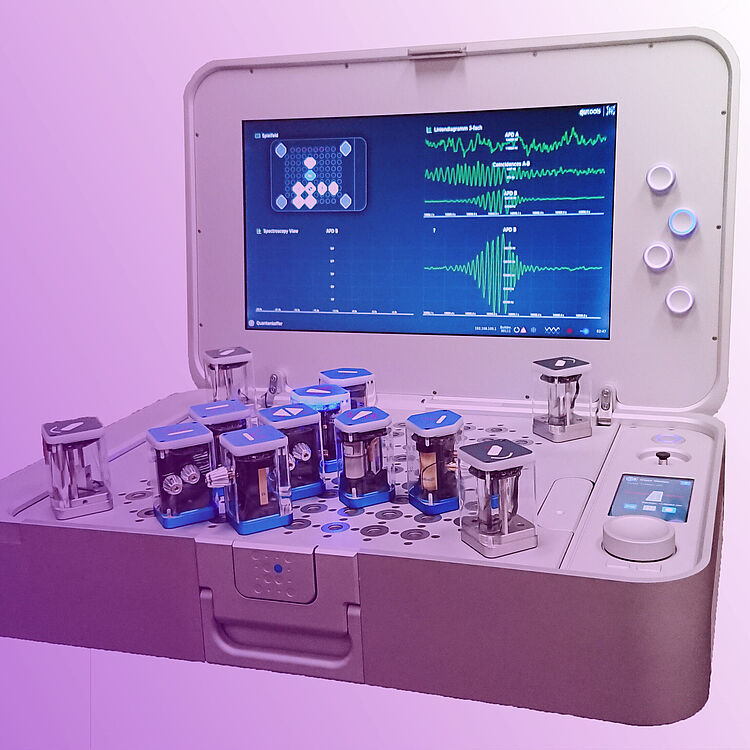 Offene weißer Quanten-Koffer mit großem Bildschirm, der grüne Wellendiagramme und Anordnungsdiagramm anzeigt, auf der Arbeitsfläche mehrere Drehregler und Tasten.