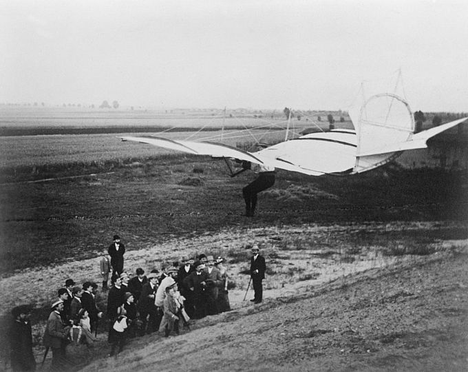 Otto Lilienthal ist noch wenige Meter vom Boden entfernt und wird mit seinem Segelapparat gleich auf einem freien Feld landen. Unten stehen circa 25 Zuschauer.