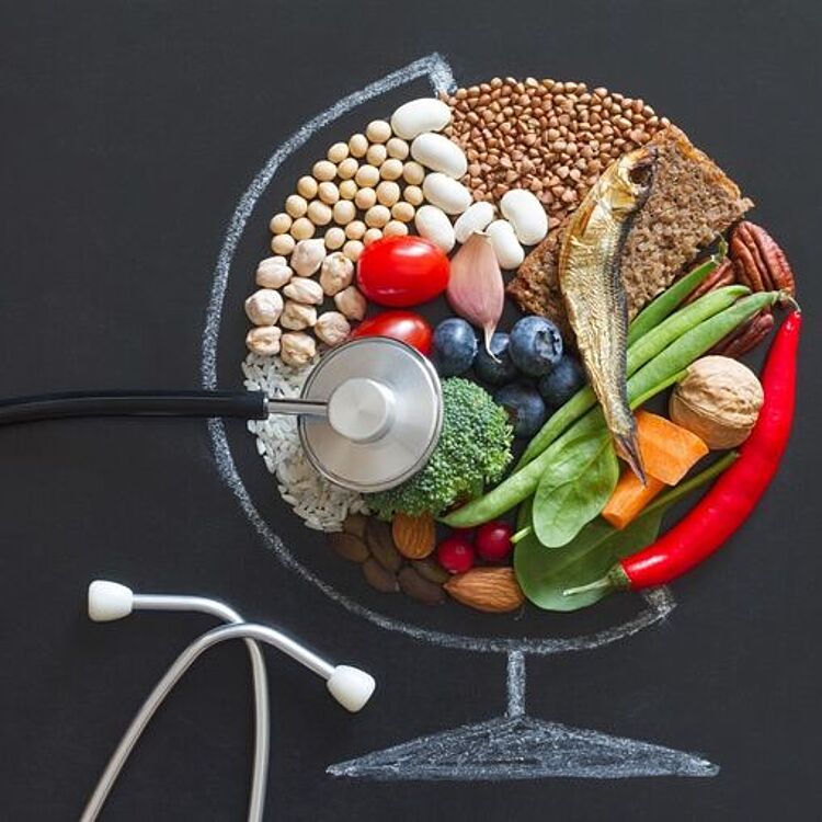 Stethoskop auf einer Tafelzeichnung eines Globus, in der die obere Hälfte mit gesunden Lebensmitteln gefüllt ist: Hülsenfrüchte, Nüsse, Reis, Vollkornbrot, Fisch, Brokkoli, Spinat, Kirschtomaten, Blaubeeren, Paprika und Chilischoten – Symbol für Ernährung und Gesundheit.