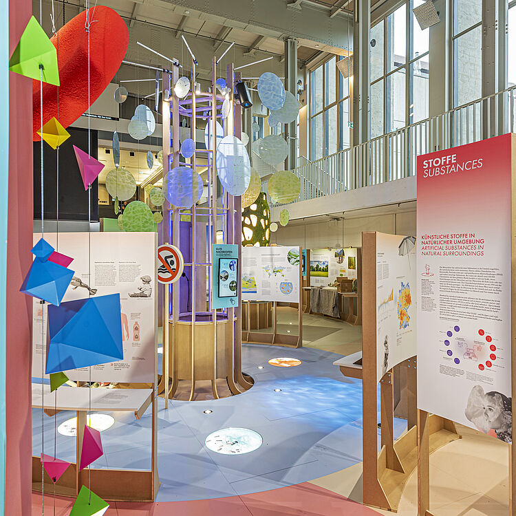 Bunte interaktive Ausstellung in einer hohen Halle mit großen Fenstern: hängende geometrische Papierformen in Pink, Grün und Blau, runde transluzente Scheiben an einer zentralen Säuleninstallation mit Lichtern, erklärende Infotafeln (rechts Titel „STOFFE SUBSTANCES“) und farbig segmentierter Boden in Rosa und Blau.