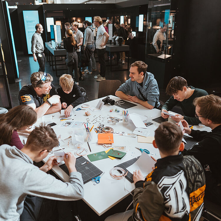 Mehrere Jugendliche sitzen an einem großen weißen Tisch in einem Workshopraum; auf dem Tisch Bastelmaterialien wie Scheren, Stifte, Papier, Klebeband, Karten und Werkzeuge verteilt, im Hintergrund weitere Besucher und Ausstellungsvitrinen in einer modernen, gut beleuchteten Ausstellungs- oder Lernumgebung.