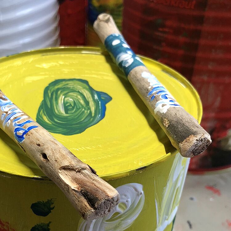 Bunt bemalte Blechdose mit zwei bemalten Holzstöcken als Trommel – ein selbst gebasteltes Musikinstrument aus einem Kinderworkshop.