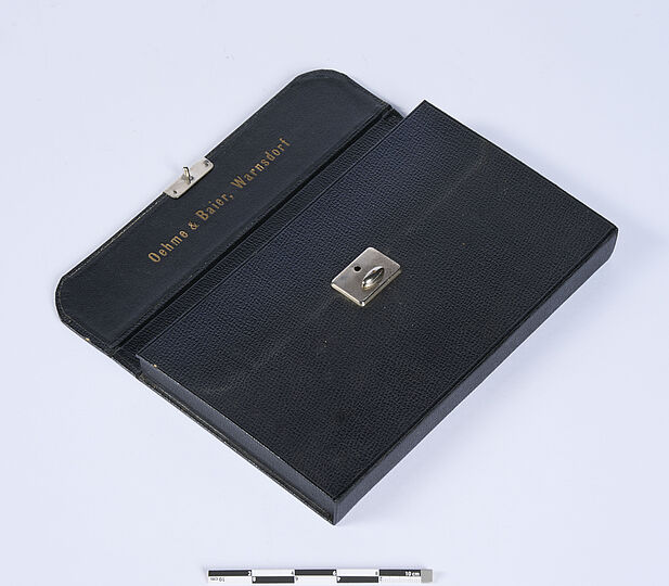 Geöffnetes, aber nicht weiter aufgeklapptes schwarzes Etui mit der Goldprägung "Oehme & Baier, Warnsdorf" im Deckel.