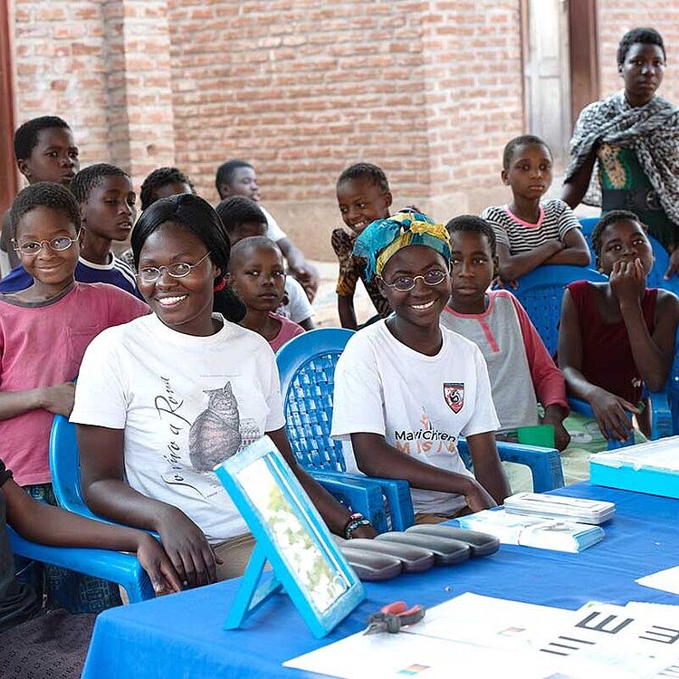 Gruppe von Kindern und Jugendlichen sitzt auf blauen Kunststoffstühlen in einem Hof vor einer Backsteinwand; im Vordergrund ein mit blauem Tuch bedeckter Tisch mit Sehtestkarten, mehreren Brillenetuis, einem kleinen Spiegel und weiteren optischen Hilfsmitteln.