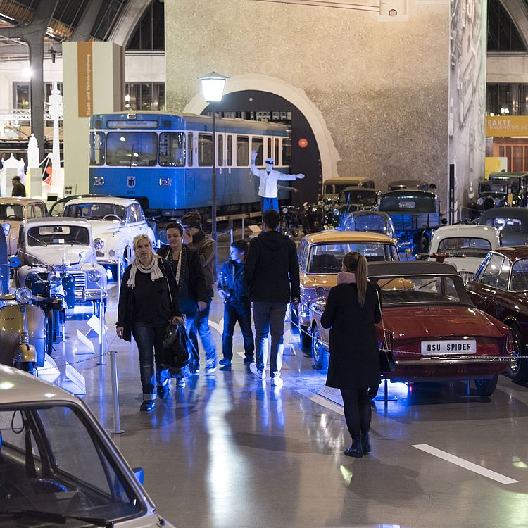 Menschen schlendern durch eine große Fahrzeugausstellung in einer Halle mit Retro-Autos und -Rollern, zwei roten NSU Spider-Cabrios im Vordergrund, links eine blaue Straßenbahn im Hintergrund, Ausstellungsinseln, Informationsschilder und sanfte blaue Beleuchtung auf dem Boden.