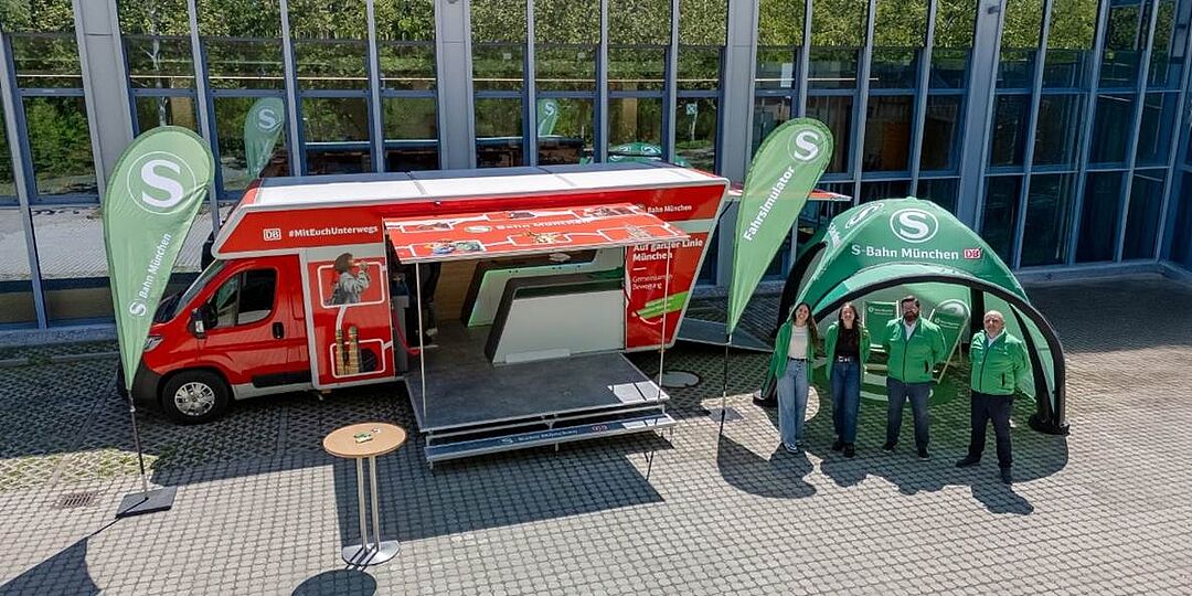 Mobiler Infostand der S-Bahn München mit grünem Pavillon und Team in grünen Jacken vor einem roten Bus mit Simulator.