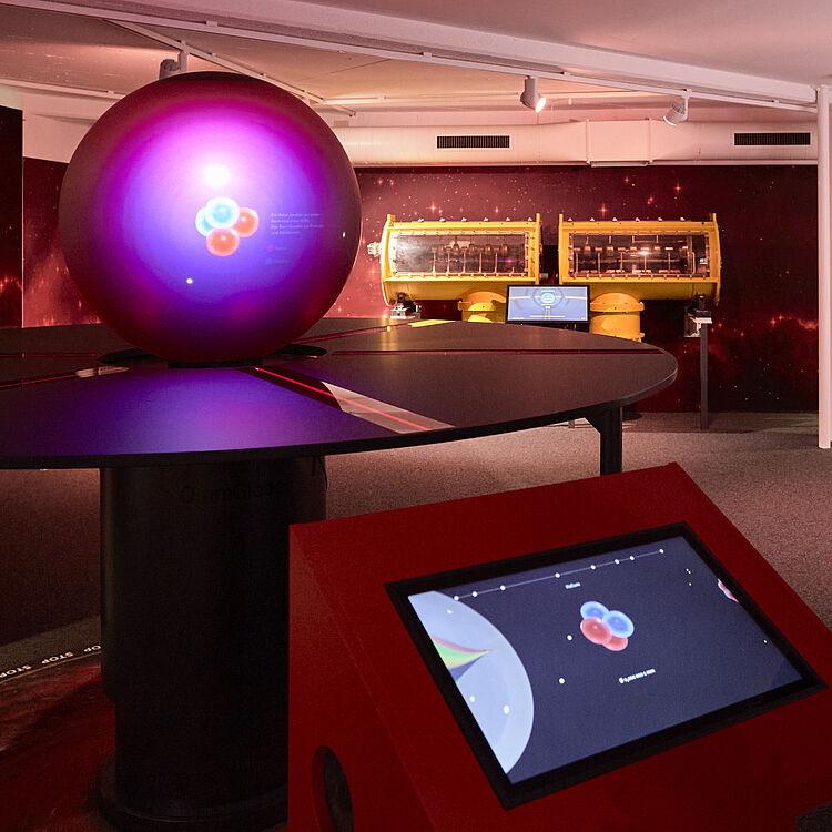 Ein Besucher betrachtet in einer dunklen Ausstellungshalle ein interaktives Wissenschafts-Exponat: eine leuchtende, violett-pinke Kugel auf einem großen schwarzen Tisch mit Projektion von Atomkern- bzw. Teilchengrafiken; im Vordergrund ein rotes Touchscreen-Display mit einer schematischen Atomdarstellung, im Hintergrund gelbe Maschinengehäuse und Sternenhimmel-Illustrationen an den Wänden.