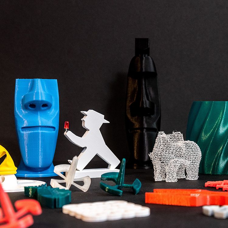 Verschieden farbige Objekte aus dem 3D-Drucker. Unter anderem drei Kreisel, einige Untersätzer, ein Berliner Ampelmännchen, eine Vase, eine kleine Nachbildung des schiefen Turm von Pisa und drei Moai-Köpfe.
