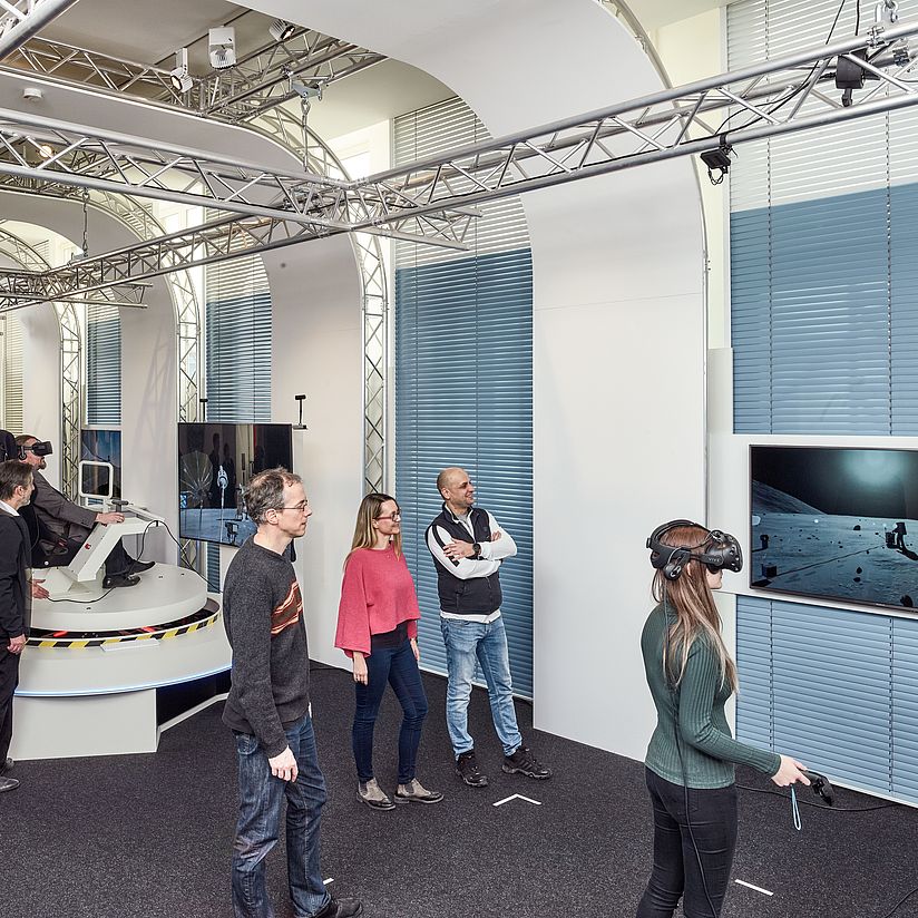 Personen im VRlab. Frau mit VR-Brille vor einem großen Monitor. Mann in einem Simulator.