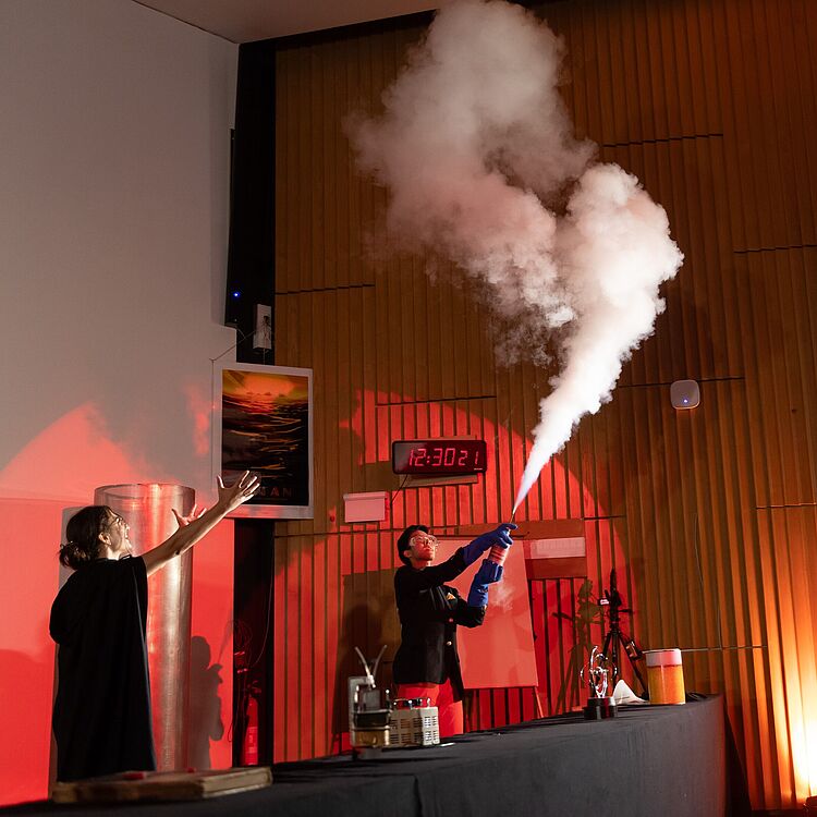 In einem Auditorium führt eine Person in schwarzem Mantel ein wissenschaftliches Experiment vor: sie hält eine blaue Druckflasche, aus der eine dicke weiße Dampfwolke nach oben schießt; links eine zweite Person mit erhobener Hand, rote Scheinwerferbeleuchtung, Tisch mit Laborutensilien im Vordergrund, digitale Uhr an Holzwand zeigt 12:30:21.