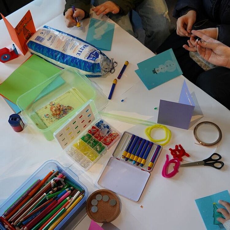 Topview: Kinder sitzen am Tisch und basteln aus Papier Karten, auf dem Tisch liegen Stifte, Watte eine Schere und Wachsmalkreide.