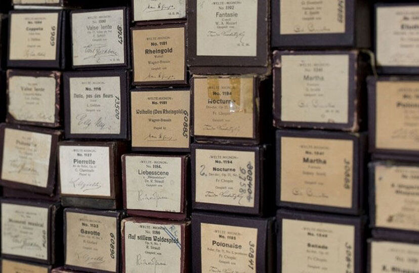 Eine Sammlung von alten Notenrollen für mechanische Musikinstrumente, aufbewahrt in dunklen Kartons mit handschriftlichen und gedruckten Etiketten. Die Etiketten zeigen Titel klassischer Musikstücke, Komponistennamen und Seriennummern. Die Rollen sind dicht gestapelt, mit sichtbaren Alters- und Gebrauchsspuren auf den Kartons.
