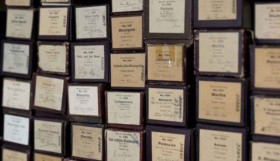 Eine Sammlung von alten Notenrollen für mechanische Musikinstrumente, aufbewahrt in dunklen Kartons mit handschriftlichen und gedruckten Etiketten. Die Etiketten zeigen Titel klassischer Musikstücke, Komponistennamen und Seriennummern. Die Rollen sind dicht gestapelt, mit sichtbaren Alters- und Gebrauchsspuren auf den Kartons.