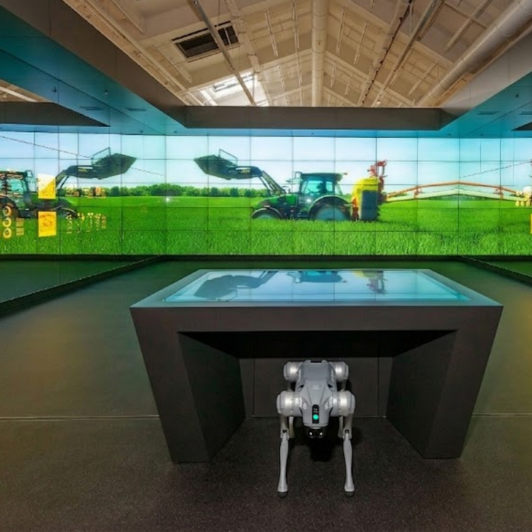 Ein grauer, mechanischer Roboterhund steht im Vordergrund unter einem digitalen Präsentationstisch. Im Hintergrund erstreckt sich eine raumhohe Video-Panoramawand, die moderne Traktoren bei der Feldarbeit zeigt.