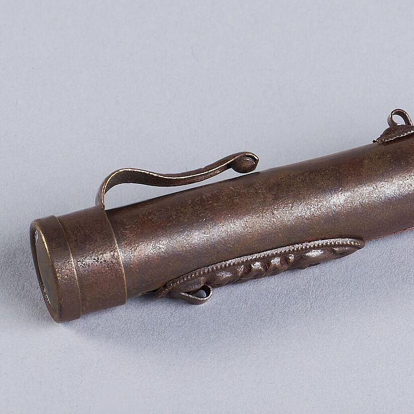 Zylindrische Messinghülle für Lippenstift liegend abgebildet. Verzierung auf der Vorder- und Halterung auf der Rückseite erkennbar. Messingdeckel an beiden Enden