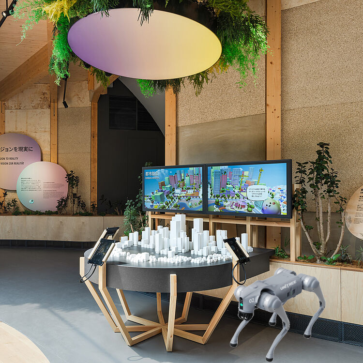Ein vierbeiniger Unitree-Roboterhund steht im Vordergrund einer hellen, mit Holz und Pflanzen gestalteten Ausstellung. Dahinter befindet sich eine interaktive Station mit einem weißen 3D-Stadtmodell, Tablets und Bildschirmen mit dem Titel Urban Mining.