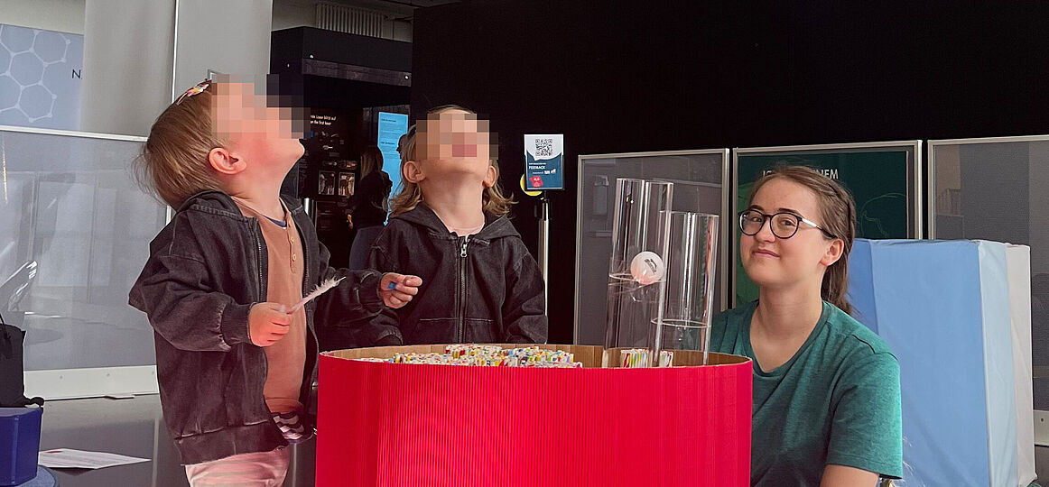 Kleinkinder schauen nach oben und spielen mit einer großen bunten Schüssel in der Strohhalme und Röhren mit Tischtennisbällen stecken.