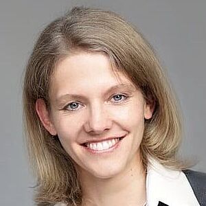 Porträt einer Frau mit schulterlangem hellblondem Haar, die einen dunklen Blazer, eine weiße Bluse und eine Halskette mit rundem Anhänger trägt; Gesicht unkenntlich gemacht.