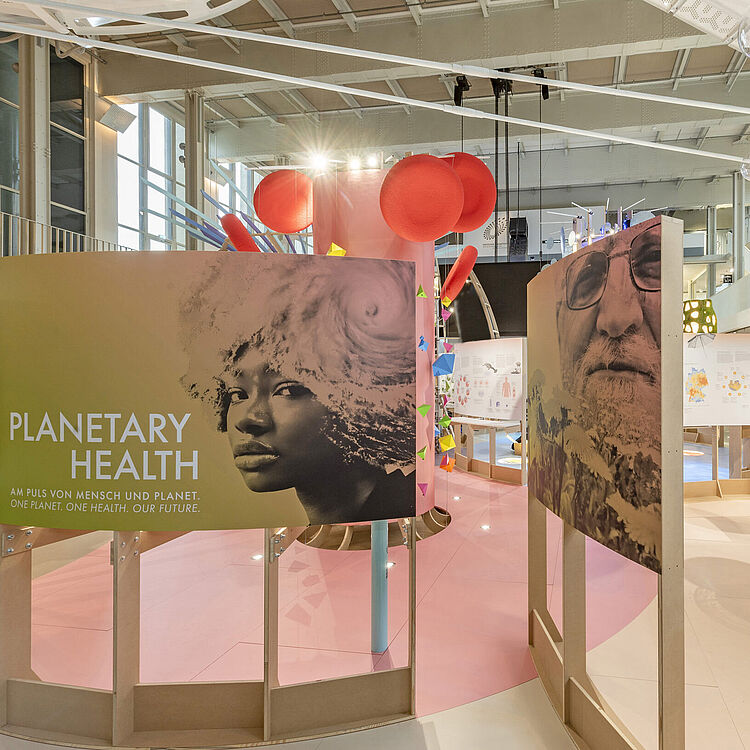 Ausstellungsraum zur „Planetary Health“-Sonderausstellung mit großem gelb-grünen Schautafel „PLANETARY HEALTH“ vorne links, runden roten Hängedekorationen oben, mehreren frei stehenden Informationstafeln mit Fotos und Texten, rechts eine blaue Infotafel mit Foto und Text, gläserne Kugelvitrinen und große silberne, scheibenförmige Hängeleuchten – modernes Museumsszenario zu Mensch-Planet-Gesundheit.
