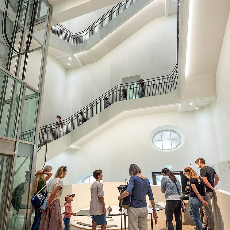 Besucher stehen im Museum vor einem Foucault’schen Pendel und beobachten dessen gleichmäßige Schwingung.