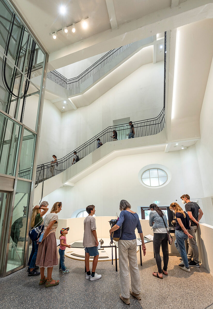 Besucher stehen im Museum vor einem Foucault’schen Pendel und beobachten dessen gleichmäßige Schwingung.