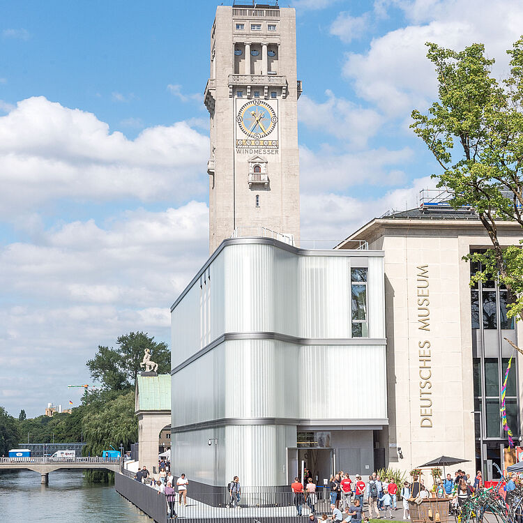 Blick auf den Turm mit großem Zifferblatt der Museumsinsel und das Gebäude des Deutschen Museums am Flussufer, moderner gläserner Anbau, Spazierweg entlang der Ufermauer, Menschen auf Bänken und Besuchern vor dem Museum, blauer Himmel mit Wolken.