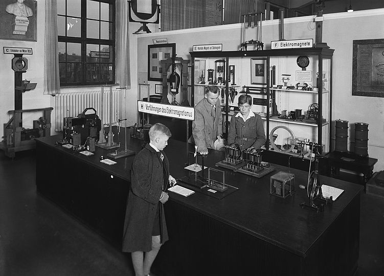 Tisch: "Vorführung des Elektromagnetismus" und Vitrinen mit "Natürliche Magnete und Stahlmagnete" - "Elektromagnete" mit drei Schülern in der Physikabteilung vor 1945.