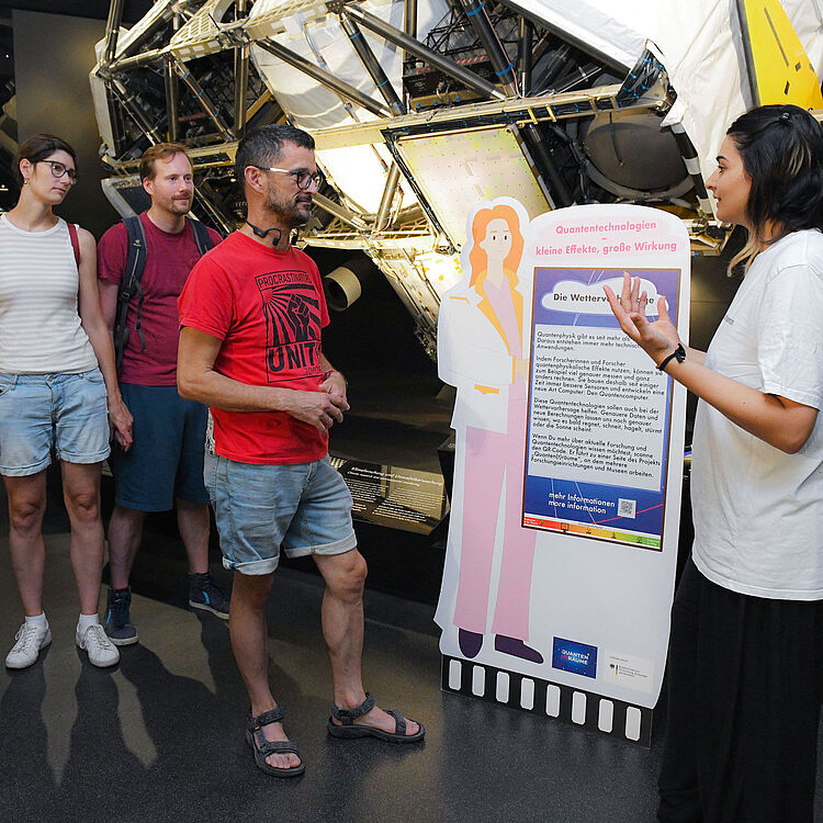 Eine Museumspädagogin in weißem T‑Shirt erklärt drei Besuchern (zwei Männer, eine Frau) vor einem Ausstellungsexponat mit technischen Metallstrukturen; vorne steht eine farbige Pappaufsteller-Infotafel mit dem Titel "Quantentechnologien" und Text.