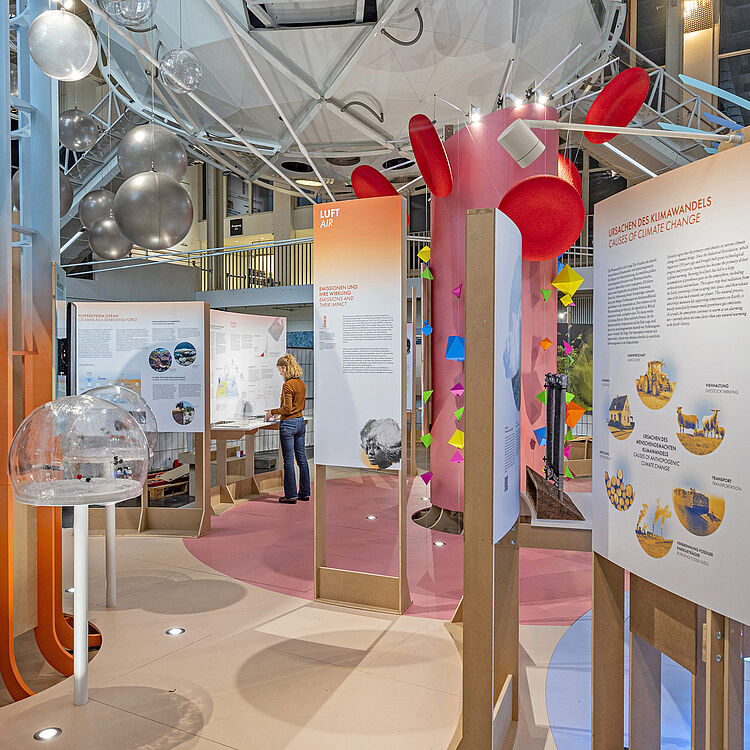 Ausstellungsraum mit weißem DNA-Besucherlabor, hängenden transparenten und grauen Kugeln, informativen Ausstellungstafeln zu Klima und Luft, farbigen Origami-Anhängern und einer Person, die vor einem interaktiven Exponat steht.