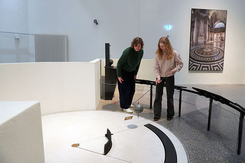 Daniela Schneevoigt und Amalia stehen am Foucaultschen Pendel im Deutschen Museum und betrachten die schwingende Kugel.