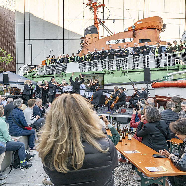 Publikum sitzt an Bierzeltgarnituren und auf Stufen, während ein Orchester und ein Chor auf der Rampe vor zwei Rettungsbooten (Schiffe beschriftet "THEODOR HEUSS" und "ASMUS BREMER") musizieren; Dirigent hebt die Arme, viele Zuschauer fotografieren mit Smartphones.