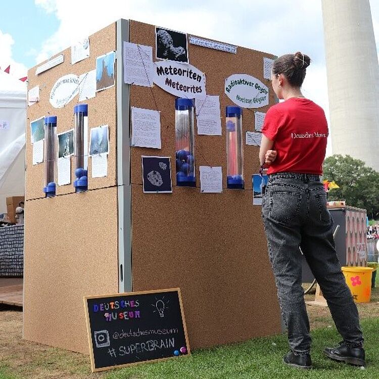 Eine Ausstellungswand im Freien mit Informationen zu Meteoriten und runden Plexiröhren, vor der eine Frau steht und die Tafeln betrachtet; im Vordergrund eine kleine schwarze Tafel mit der Handlettering-Aufschrift „Deutsches Museum“ und dem Hashtag „SUPERBRAIN“.