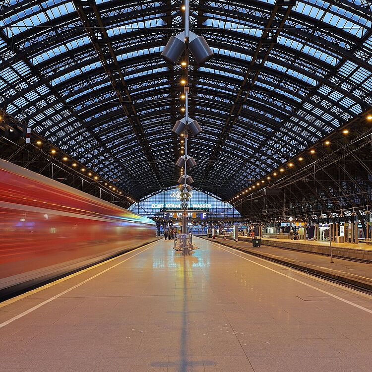 Die Bahnsteighalle des Kölner Hauptbahnhofs bei Abendstimmung. Das Abendlicht fällt durch die filigrane Stahlkonstruktion des halbrund gewölbten Hallendachs hoch über den Bahnsteigen.