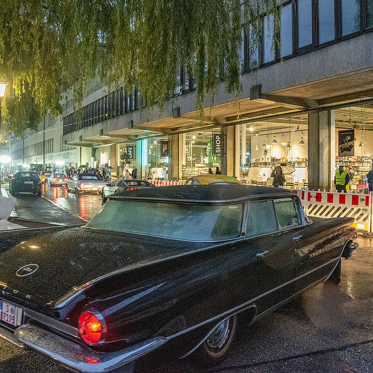 Schwarzes Vintage-Auto (Knudsen/Oldtimer) mit roten Rücklichtern auf nasser Straße vor einem beleuchteten Laden- und Cafégebäude bei Regen, Menschen, Absperrungen und Bäumen mit herabhängenden Zweigen im Abendlicht.