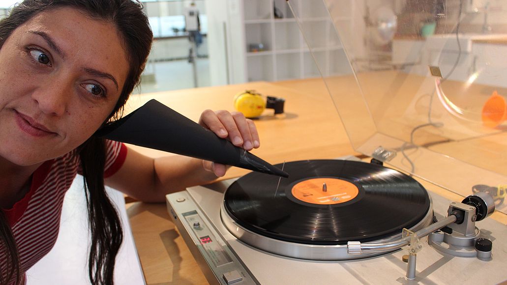 Frau hört mit einem Trichter aus Pappe eine Schallplatte. Der Trichter hat am Ende eine Nadel. Diese gleitet über die Schallplatte, während sie sich dreht.