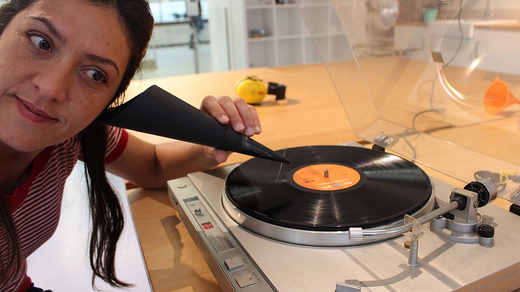 Hör-Werkstatt Frau hört mit einem Trichter aus Pappe eine Schallplatte. Der Trichter hat am Ende eine Nadel. Diese gleitet über die Schallplatte, während sie sich dreht.