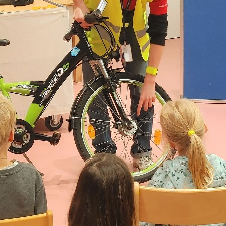 Eine Person in einer gelben Warnweste zeigt einer Gruppe von Kindern ein Fahrrad. Der Fokus liegt auf den Reifen und reflektierenden Elementen des Fahrrads, um das Thema Licht und Reflexion zu erklären. Die Kinder sitzen auf Stühlen und beobachten aufmerksam.