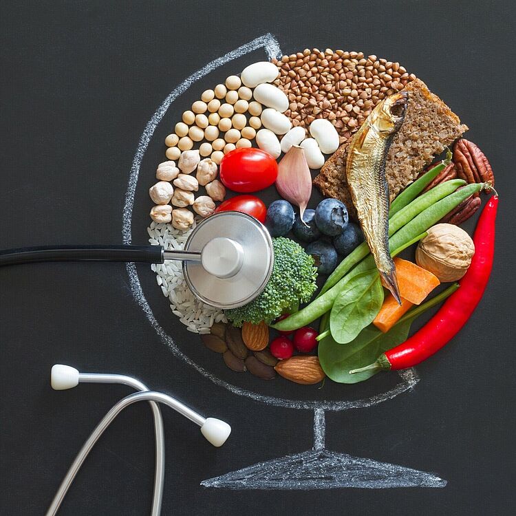 Stethoskop auf einer Tafelzeichnung eines Globus, dessen oberes Viertel mit gesunden Lebensmitteln gefüllt ist: Fisch, Vollkornbrot, Bohnen, Linsen, Reis, Nüsse, Mandeln, Brokkoli, Spinat, Tomaten, Blaubeeren, Chili und andere Gemüse.