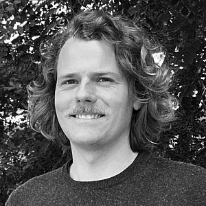 Schwarzweiß-Porträt einer Person mit geschulterlangem, lockigem Haar, trägt einen dunklen Rundhalsausschnitt-Pullover; Gesicht anonym durch graues Rechteck verdeckt, Hintergrund dichter Laubbewuchs.