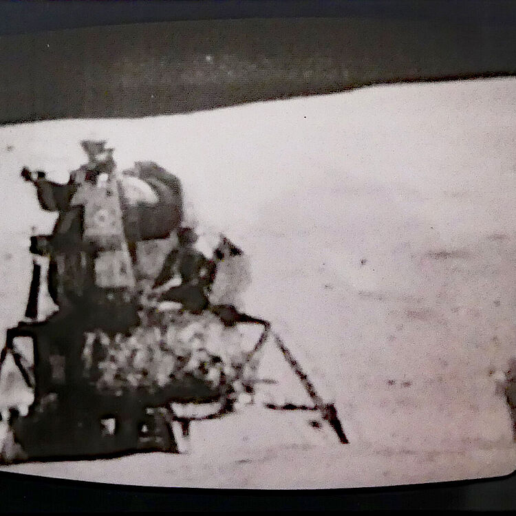 Schwarzweißaufnahme der Apollo-Landefähre auf der Mondoberfläche, links die Abstiegstreppe und Landebeine, rechts weite, leicht strukturierte Mondlandschaft.