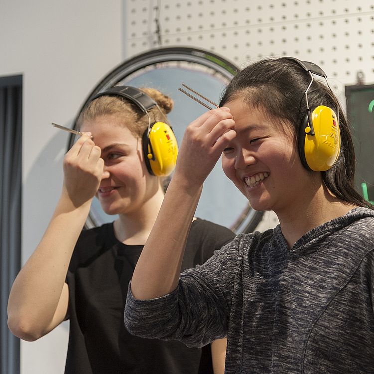 Zwei Besucherinnen in der Experimentier-Werkstatt. Sie tragen beide einen Hörschutz und halten sich eine Stimmgabel an die Stirn.