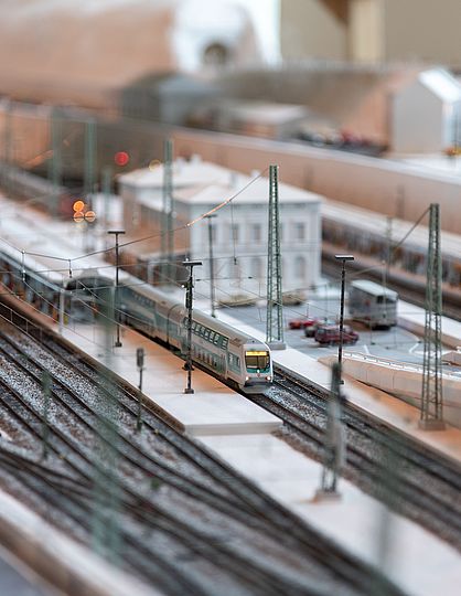 Detail der Modelleisenbahn. Zu sehen sind der Bahnhof mit Parkplatz, zwei überdachte Bahnsteige und ein einfahrender Zug.