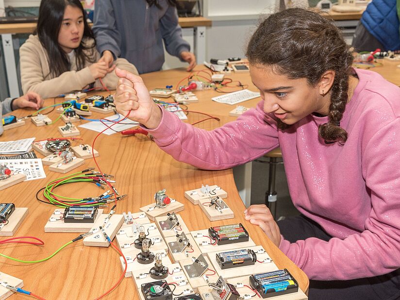 Ein Mädchen beschäftigt sich mit Kabeln und Steckverbindungen eines Schaltkreises (vor ihr auf dem Tisch aufgebaut).