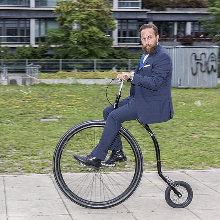 Ein Mann in einem dunkelblauen Anzug und schwarzen Lederschuhen fährt ein hochradähnliches Fahrrad (Penny-Farthing-Stil) auf einem Bürgersteig vor einer Rasenfläche und modernen Gebäuden; Gesicht absichtlich verpixelt.